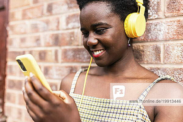 Smiling woman using smart phone and listening to music in front of brick wall