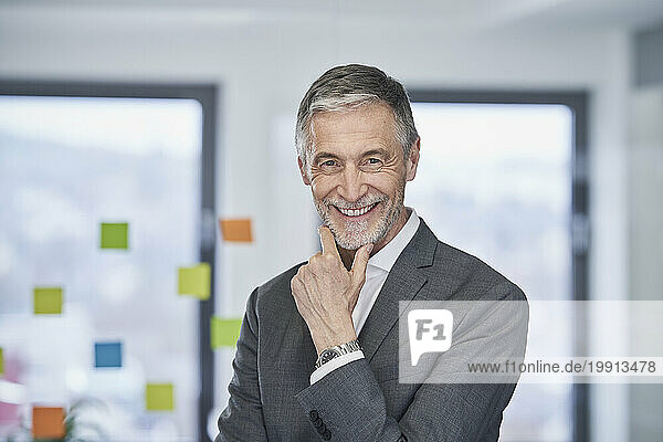 Happy senior businessman with hand on chin standing in office