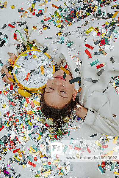 Happy girl holding clock and lying on confetti