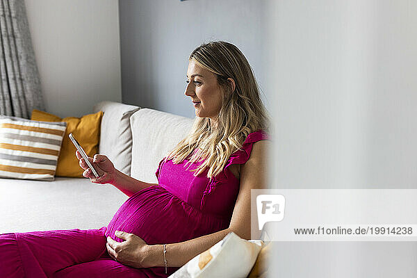 Smiling pregnant woman sitting on sofa and using smart phone at home