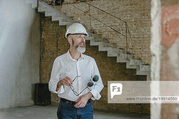 Happy architect standing with eyeglasses and documents at construction site