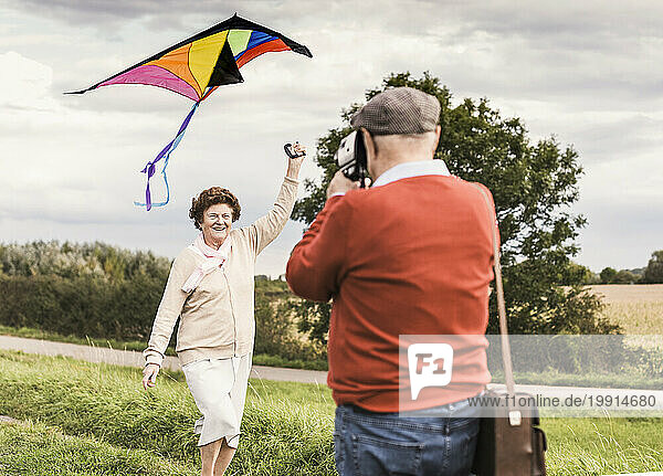 Senior man taking picture of woman flying kite at field
