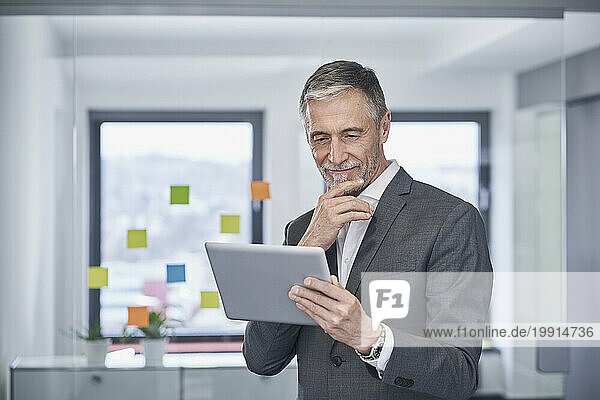 Smiling senior businessman with hand on chin using tablet PC in office