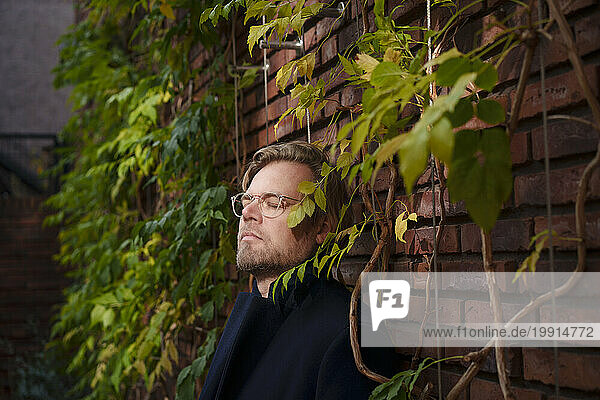 Businessman standing with eyes closed leaning on brick walli overgrown with plants
