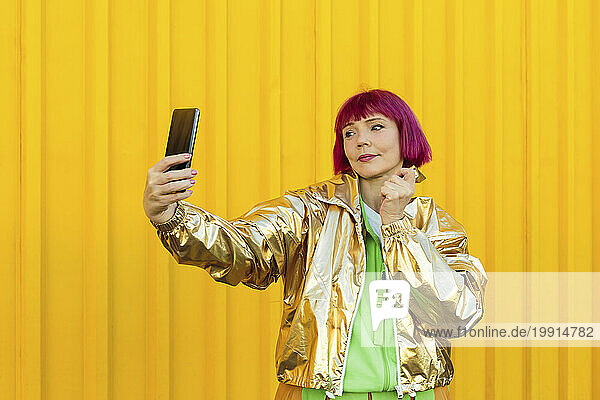 Smiling senior woman taking selfie with smart phone in front of yellow wall