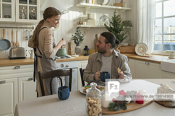 Smiling woman stirring pan and talking to man sitting at table