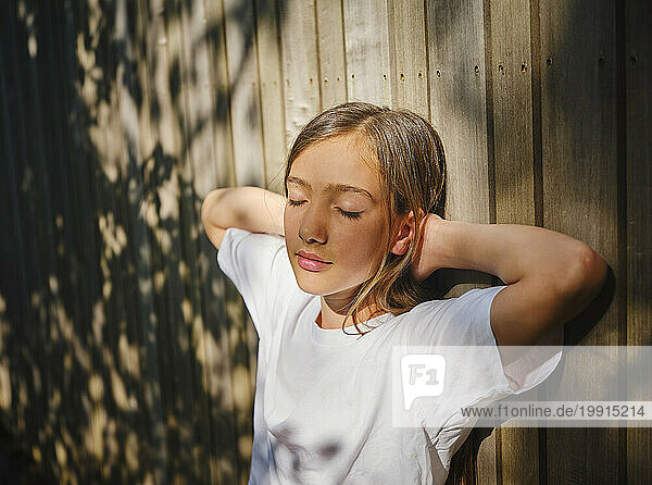 Girl with eyes closed leaning on wooden wall and enjoying sunlight