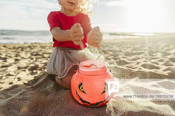 Girl filling sand in Halloween pumpkin toy at beach on sunny day