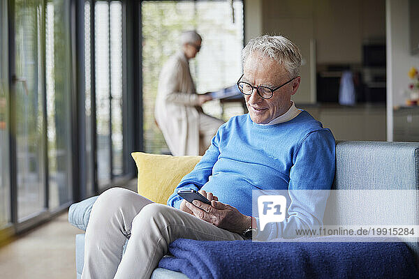 Senior man sitting on couch at home using mobile phone