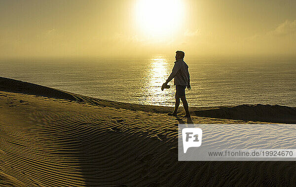 Man walks along sand dune with sunset and ocean in background.