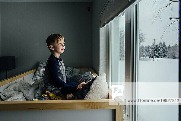Bright side view of little boy sitting on bed looking out window