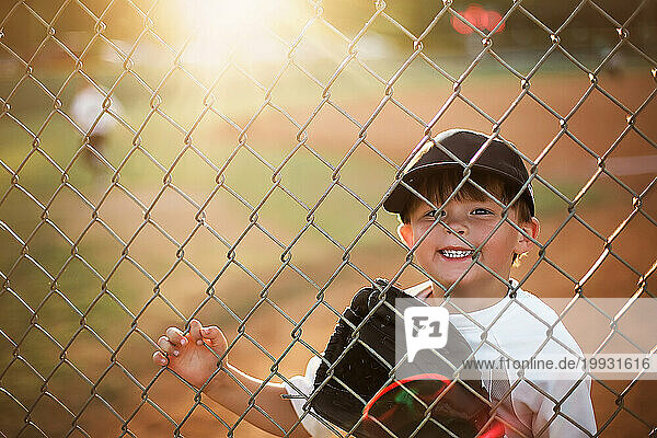 Little boy at a baseball game looking through the fence smiling