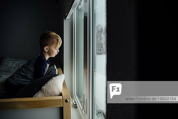 Small boy sits on bed and looks out window at snow below with dr