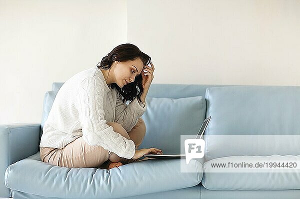 Junge nachdenkliche Frau in Freizeitkleidung  die mit den Beinen auf dem Sofa sitzt und die Tastatur des Laptops benutzt  auf den Computerbildschirm starrt und nach wichtigen notwendigen Informationen sucht oder dringende E Mails beantwortet