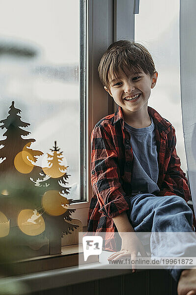 Smiling boy sitting on window sill near Christmas decoration at home