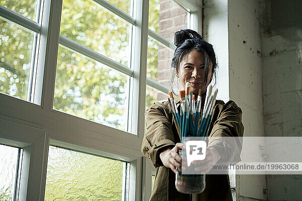 Smiling painter holding paintbrushes standing by window at workshop