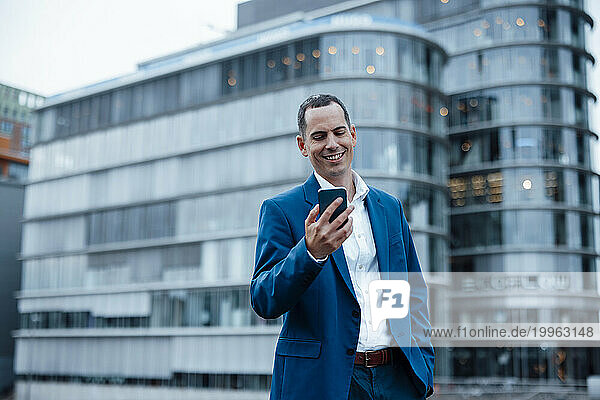 Happy businessman using smart phone near buildings