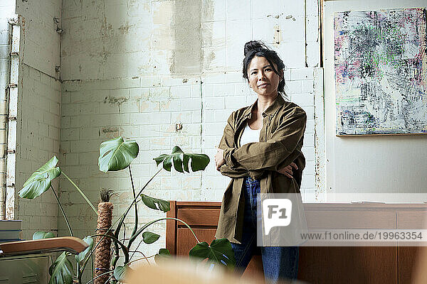 Confident artist standing with arms crossed in front of painting on wall in workshop