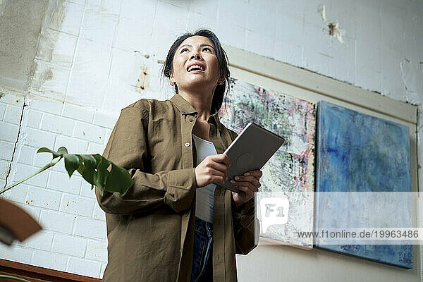 Smiling woman holding tablet PC in front of paintings at art studio