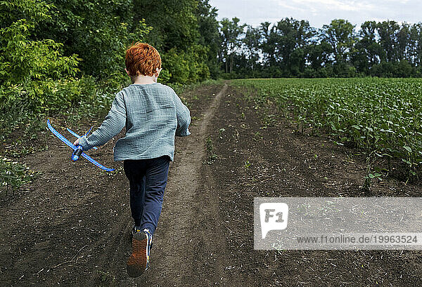 Redhead boy holding toy airplane and running on dirt road near field
