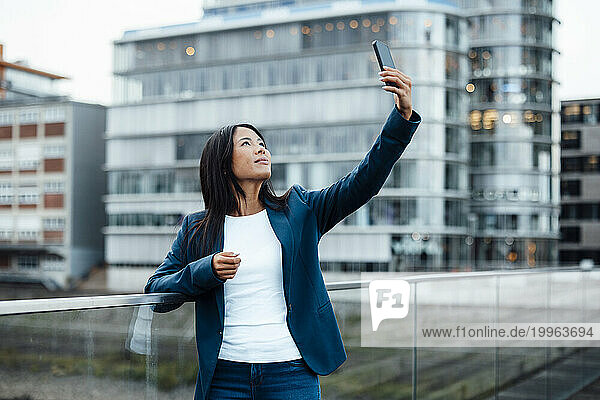 Smiling businesswoman taking selfie near railing in front of buildings