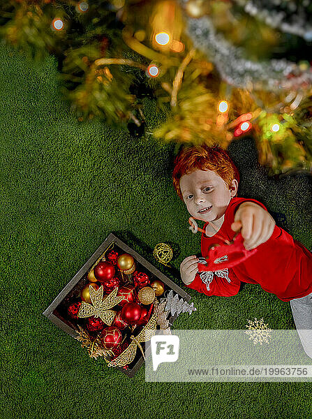 Happy redhead boy playing with ornament by Christmas tree on green carpet