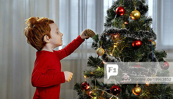 Redhead boy decorating Christmas tree with pine cone at home