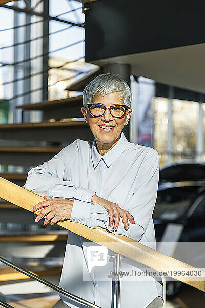 Smiling businesswoman wearing eyeglasses and leaning on railing at office