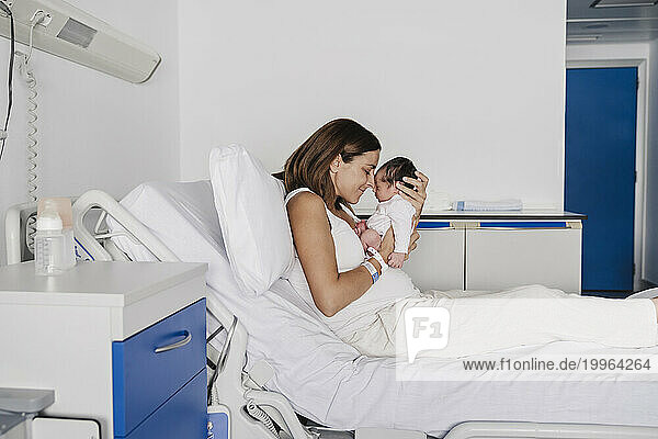 Mother sitting on bed and embracing baby daughter in hospital