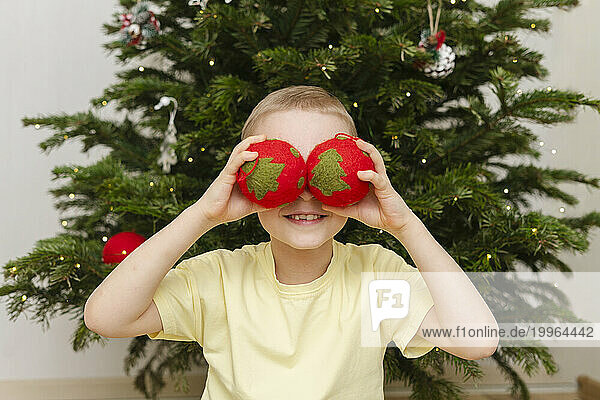 Smiling boy hiding eyes with Christmas ornaments at home