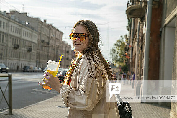 Smiling young woman holding glass of juice on sidewalk in city