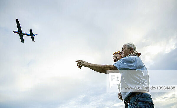 Smiling grandfather carrying grandson and flying toy airplane under cloudy sky