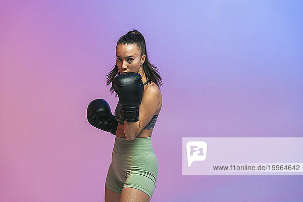 Confident young woman exercising with black boxing gloves against colored background