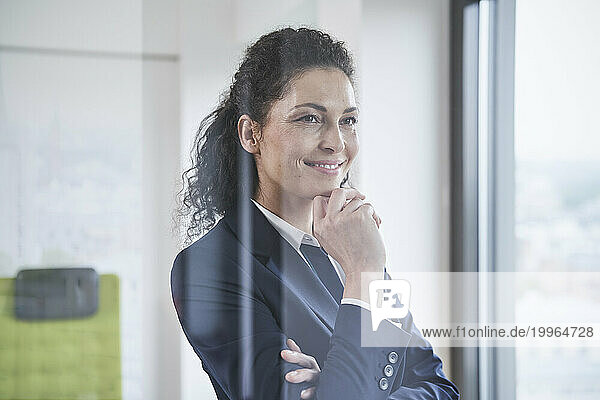 Smiling mature businesswoman with hand on chin seen through glass