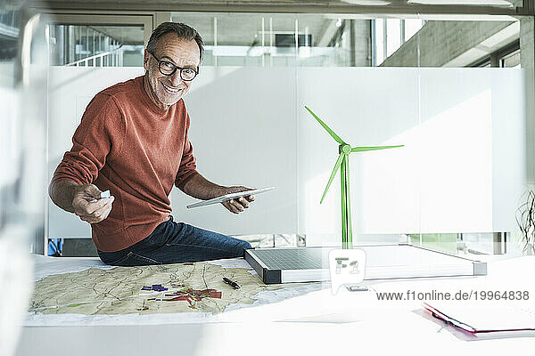 Happy businessman sitting with tablet PC near wind turbine model and solar panel on desk