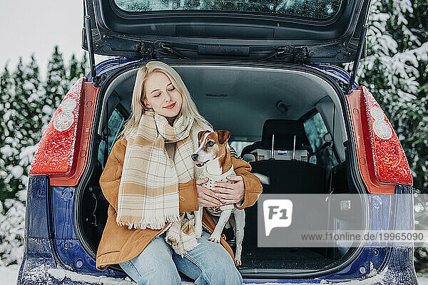 Smiling woman sitting in car trunk with pet dog