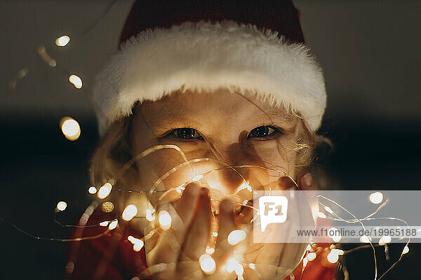 Happy girl wearing Santa hat and holding string lights in hand