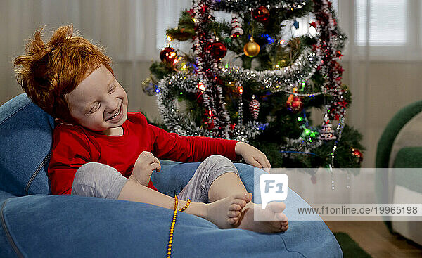 Cheerful redhead boy sitting on blue bean bag chair near Christmas tree at home