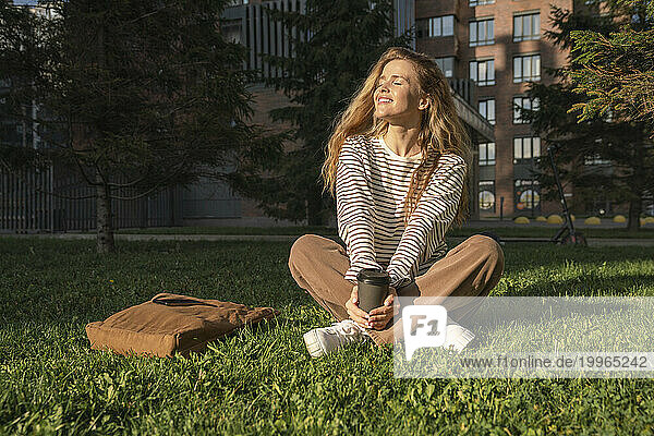 Woman with eyes closed and disposable cup sitting on grass in park