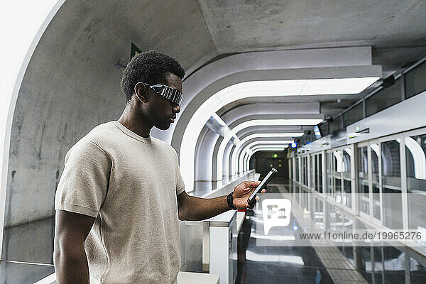 Man wearing futuristic glasses standing at metro station using smartphone