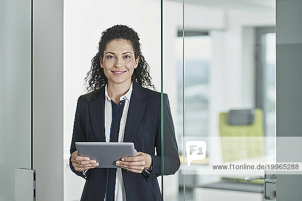 Smiling mature businesswoman standing with tablet PC in office doorway