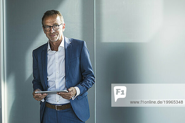 Smiling businessman standing with tablet PC in front of wall