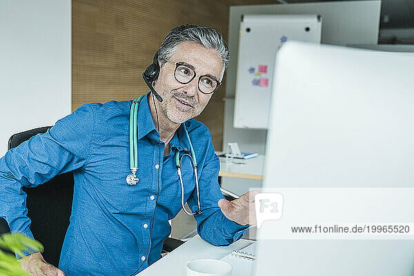 Mature doctor wearing headset and talking on video call through computer