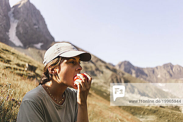 Woman wearing cap and eating tomato in front of mountains on sunny day