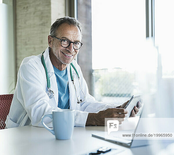 Happy doctor sitting with tablet PC and laptop at desk