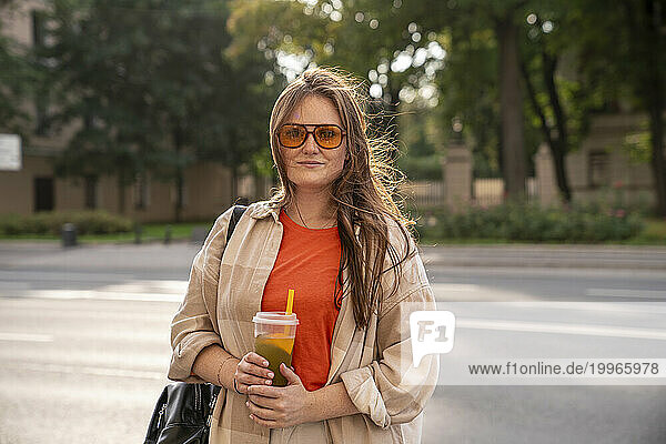 Smiling young woman holding glass of juice and standing on street