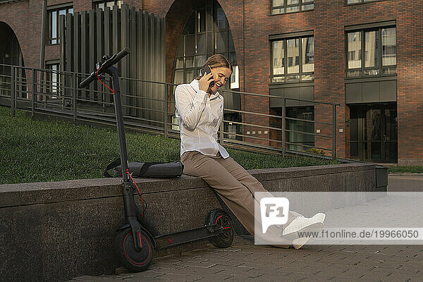 Businesswoman talking over mobile phone sitting by electric push scooter