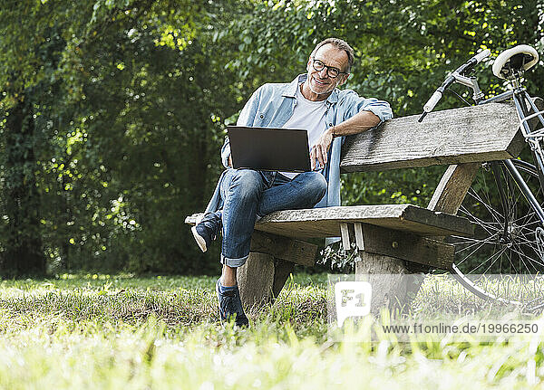Smiling man with laptop sitting on bench in park