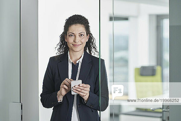 Smiling businesswoman standing with mobile phone in office doorway