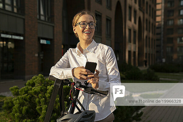 Smiling businesswoman with mobile phone and electric push scooter on footpath